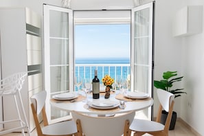 A dining table set for four with a bottle of wine and a vase of flowers, overlooking the sea.