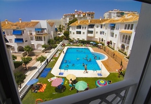 A swimming pool surrounded by white buildings with balconies, a grassy area with umbrellas, and a clear blue sky.