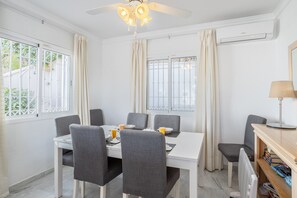 Een eetruimte met een witte tafel, grijze stoelen, een plafondventilator en een raam met gordijnen.