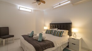 Une chambre à coucher comprenant un lit, une chaise, une table de chevet avec une lampe, un ventilateur de plafond et une fenêtre.