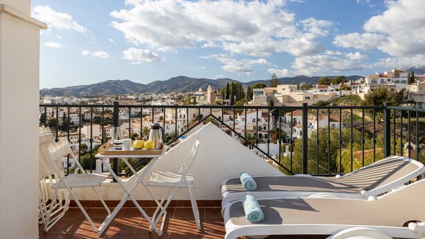 Un balcon avec une table, des chaises et des fauteuils, offrant une vue sur le paysage urbain et les montagnes au loin.