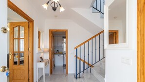 Un couloir avec une porte en bois, un bureau blanc, un escalier avec des rampes bleues et, au fond, une cuisine.