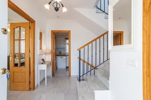 Un pasillo con una puerta de madera, un escritorio blanco, una escalera con barandillas azules y una cocina al fondo.