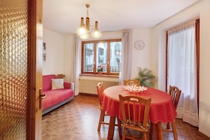 A dining area with a round table, chairs, a red tablecloth, a chandelier, a window with curtains, a radiator, and a sofa.