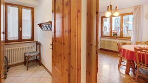 Une pièce avec une porte en bois, un radiateur, une chaise, une table recouverte d’une nappe et une fenêtre donnant sur des immeubles.