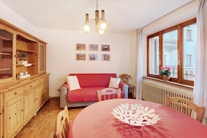 A cozy room with a wooden cabinet, a red sofa, a dining table with a white table runner, and a window with a view of buildings.