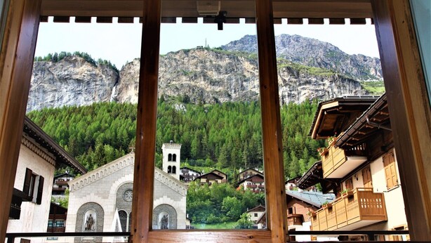 Une vue à travers la fenêtre d’un bâtiment traditionnel, doté d’une arche en pierre, de balcons en bois et d’un paysage montagneux en arrière-plan.