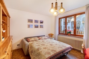 A bedroom with a wooden dresser, a bed with floral bedding, a window with a view of buildings, and a ceiling light fixture.