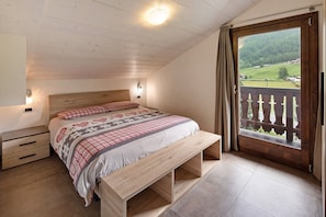 A bedroom with a wooden ceiling, a bed with a checkered bedspread, a bench, a nightstand, and a view of a balcony with a wooden railing.