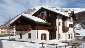 Un chalet de dos pisos con balcones de madera, una pista de esquí al fondo y un cielo despejado.