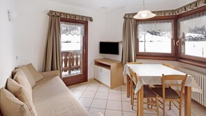 Una habitación con sofá, una mesa de comedor con sillas, televisor y ventanas amplias con cortinas.