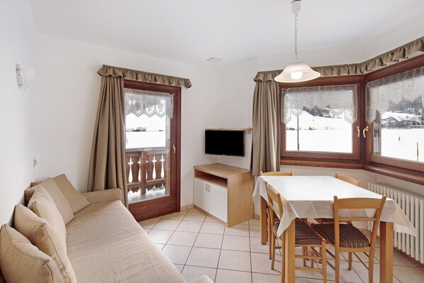 A room with a sofa, a dining table with chairs, a TV, and large windows with curtains.