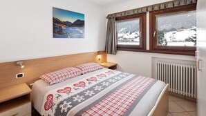 Un dormitorio con cama, mesita de noche, lámpara de pared, ventana con cortinas y un cuadro de un paisaje nevado.