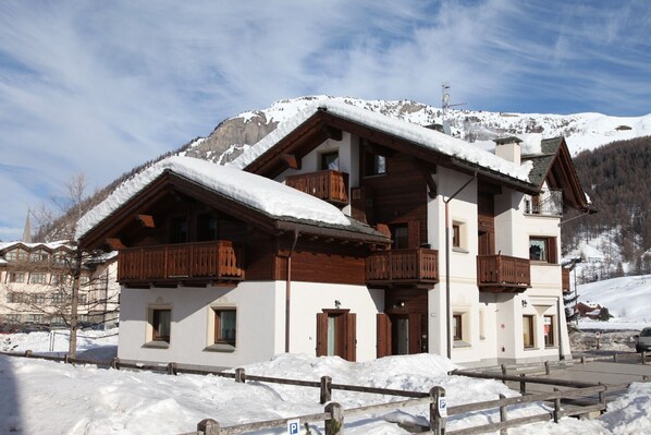 Un chalet à deux étages avec des balcons en bois, une piste de ski en arrière-plan et un ciel dégagé.