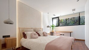 Un dormitorio moderno con cama, mesitas de noche, un escritorio y un amplio ventanal con vistas a zonas verdes.