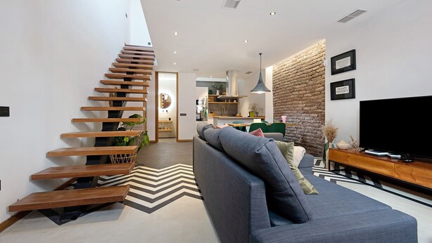 Una sala moderna con un sofá gris, una escalera de madera, una cocina al fondo y una pared de ladrillo.