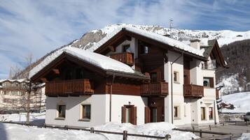 Un chalet a due piani con balconi in legno, una pista da sci sullo sfondo e cielo sereno.