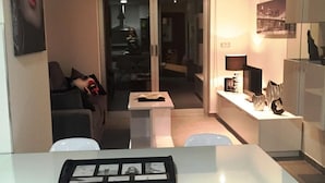 En moderne stue med glasvæg, et spiseområde med et hvidt bord, og et køkken med skabe og fjernsyn.
