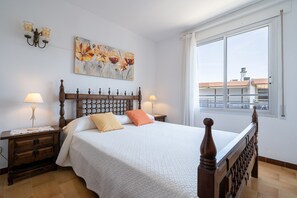 A bedroom with a large bed, two bedside tables, a wall-mounted lamp, and a window with a view of buildings.