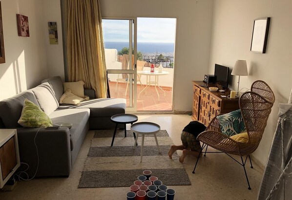 A living room with a sofa, two small tables, a wicker chair, a television, and a view of the city through a large window.