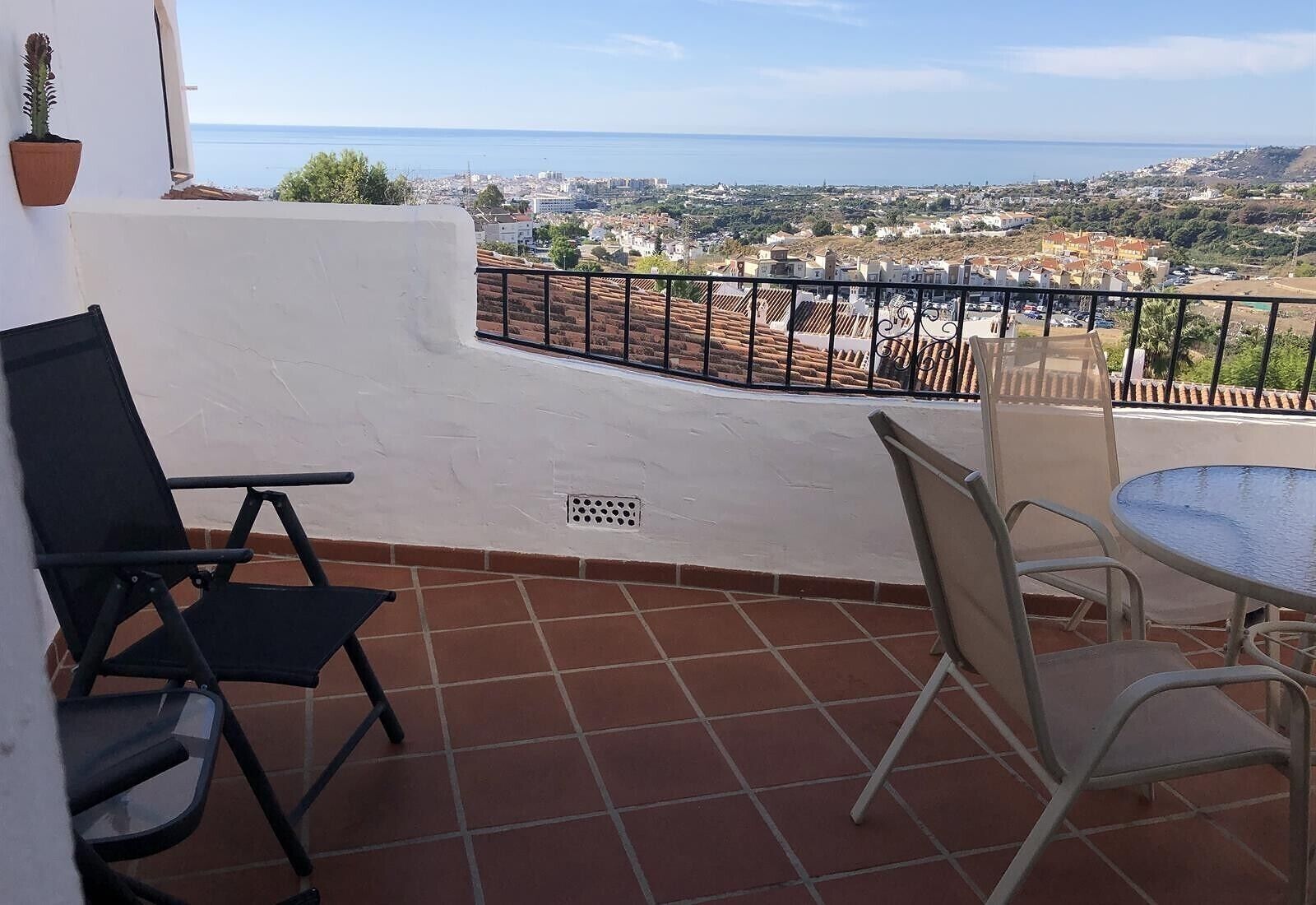 Un balcon avec vue sur la mer et la ville, deux chaises, une table et un pot de fleurs.