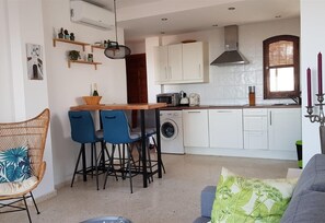 Una cocina compacta con lavadora, microondas y pileta. Hay dos sillas azules junto a una mesita, y una silla de mimbre con cojín.
