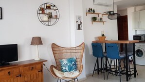 Un espace de vie compact comprenant un téléviseur, un fauteuil en osier, une table à manger avec des chaises et un lave-linge.