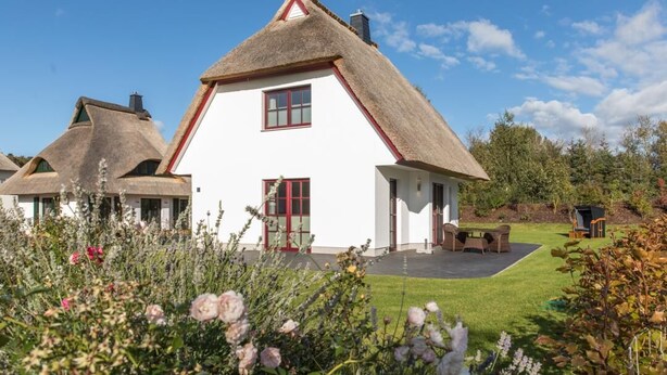 A white house with a thatched roof, surrounded by a well-maintained garden with various plants and flowers. - House Rose, luxury vacation home in Fuhlendorf for 4 people with sauna and beach chair (Fuhlendorf)