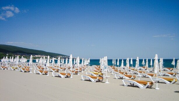 A beach with rows of sun loungers and umbrellas, a clear sky, and a distant view of a town. - Seaside Apartment with 2 Bedrooms for up to 5 Guests (Sunny Beach)