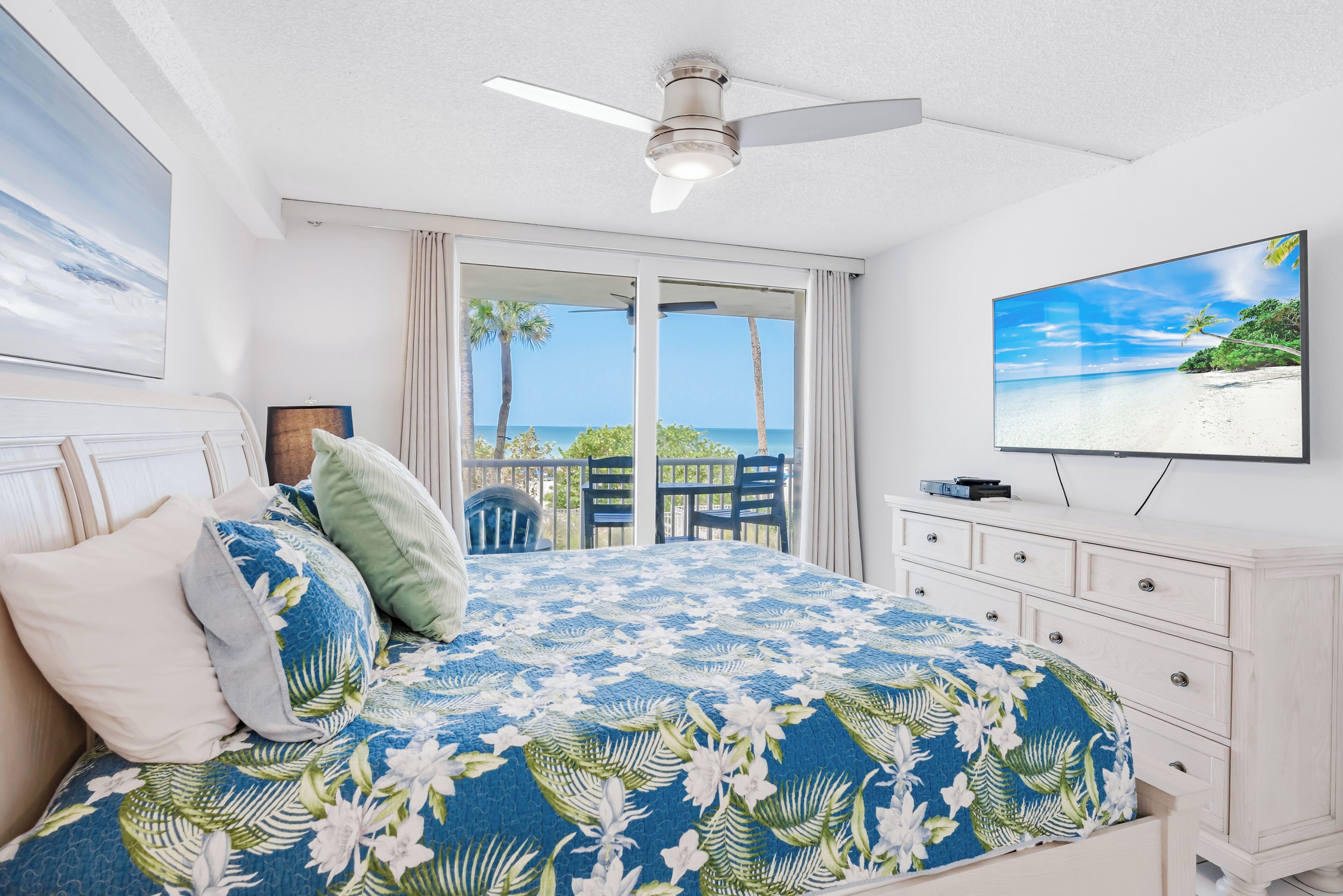 A bedroom with a bed, a ceiling fan, a TV, and a balcony with a view of the beach.