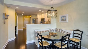 A dining area with a round table, four chairs, a chandelier, a kitchen in the background, and a door leading to another room.