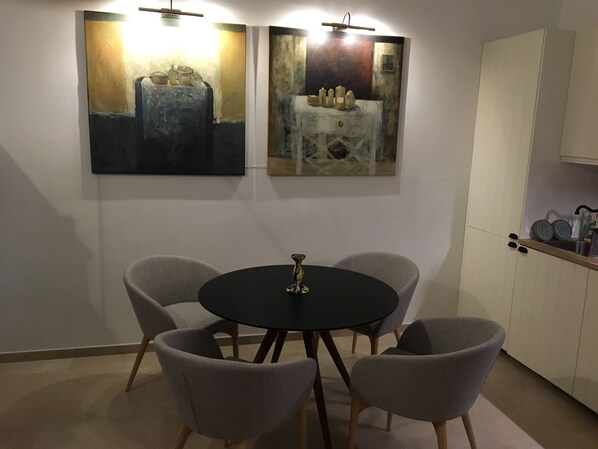 A dining area with a round table and four chairs, two paintings on the wall, and a kitchen area with cabinets and a sink. - Charming 2-Bedroom House - Rooftop terrace in Heart of Noto, Sicily (Noto)