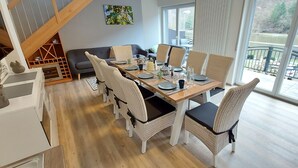 A dining area with a wooden table set for six, surrounded by wicker chairs with black cushions. A grey sofa is against the wall, and a staircase is visible in the background.