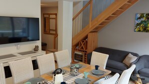 A modern living room with a flat-screen TV, a dining area with wicker chairs, a grey sofa, and a wooden staircase.
