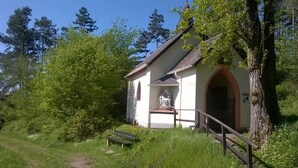A small chapel with a statue on the porch, surrounded by greenery and trees.