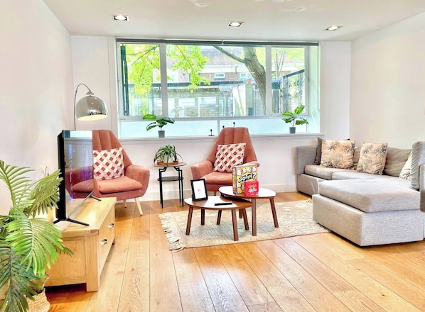 Una sala de estar moderna con un sofá, dos sillones, una mesa de centro y un televisor. Hay plantas y una ventana con vistas al exterior.