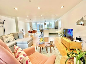 Una sala de estar moderna con sofá, mesa de centro y televisor. Se observa un área de cocina al fondo con una mesa de comedor y sillas.