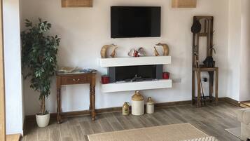 Una habitación con techo de madera, un televisor de pantalla plana sobre la chimenea, una mesa de madera con una planta y un otomano estampado.