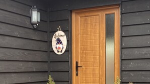 Une porte en bois ornée d’une pancarte « Bienvenue », deux plantes en pot et une lanterne fixée à un mur en bois sombre.