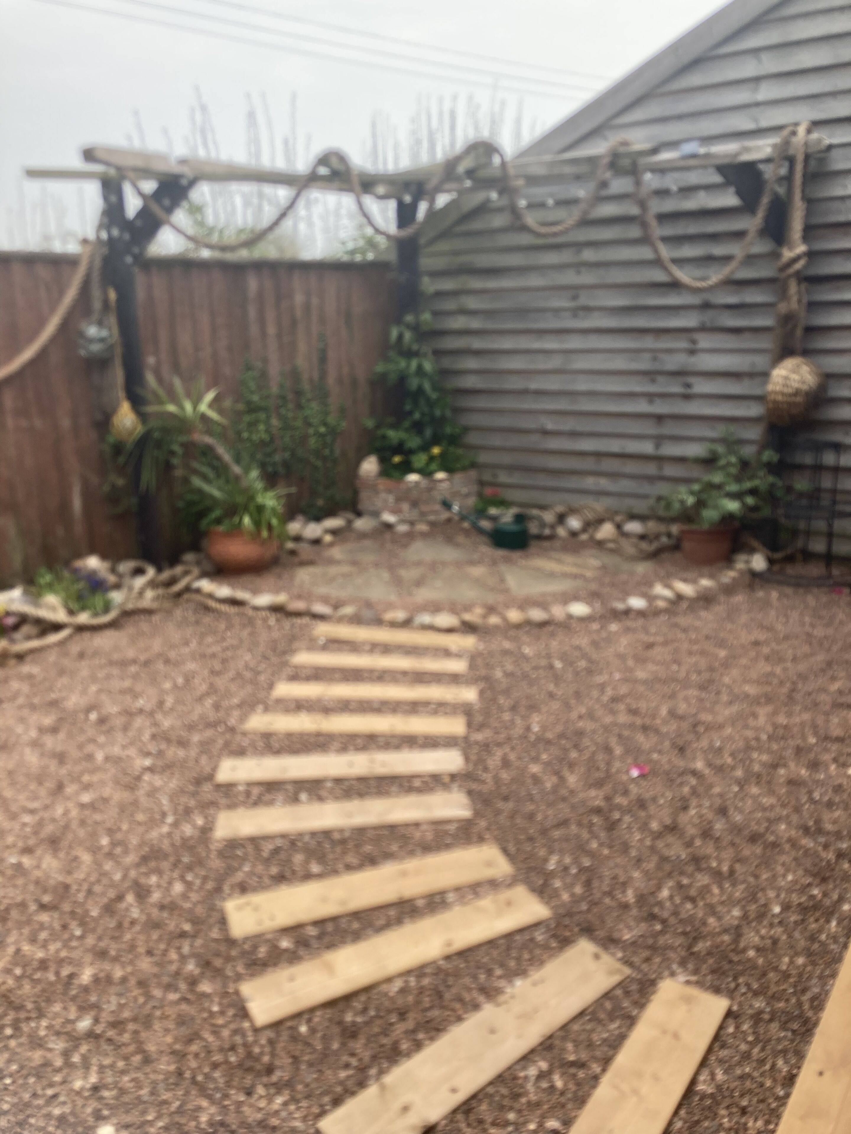 Un jardin avec un sentier en bois menant à une maison en bois. On y trouve des plantes en pot et une clôture qui entoure le jardin.