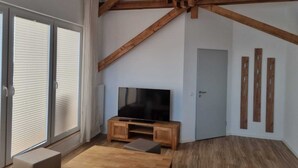 Een moderne woonkamer met zichtbare houten balken, een grote flatscreentelevisie op een houten standaard, een schuifpui en een klein houten tafeltje.