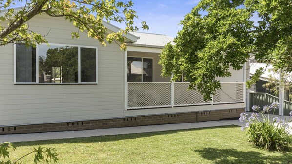 A single-story house with a white exterior, a covered porch, and a small front yard with grass and a few plants. - Coastal Soul - Belle Escapes Jervis Bay (Sanctuary Point)