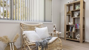 A wicker chair with cushions, a small round table with a decorative item, and a wooden bookshelf with books and decorative objects. - Coastal Soul - Belle Escapes Jervis Bay (Sanctuary Point)