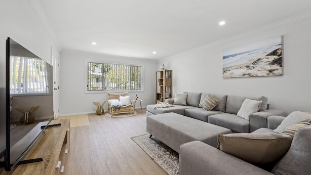 A modern living room with a grey sofa set, a flat-screen TV, and a large window with vertical blinds. - Coastal Soul - Belle Escapes Jervis Bay (Sanctuary Point)