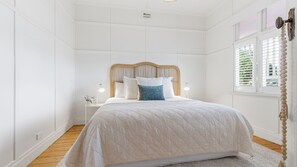 A bedroom with a bed, bedside lamps, a window with blinds, and a wooden floor.