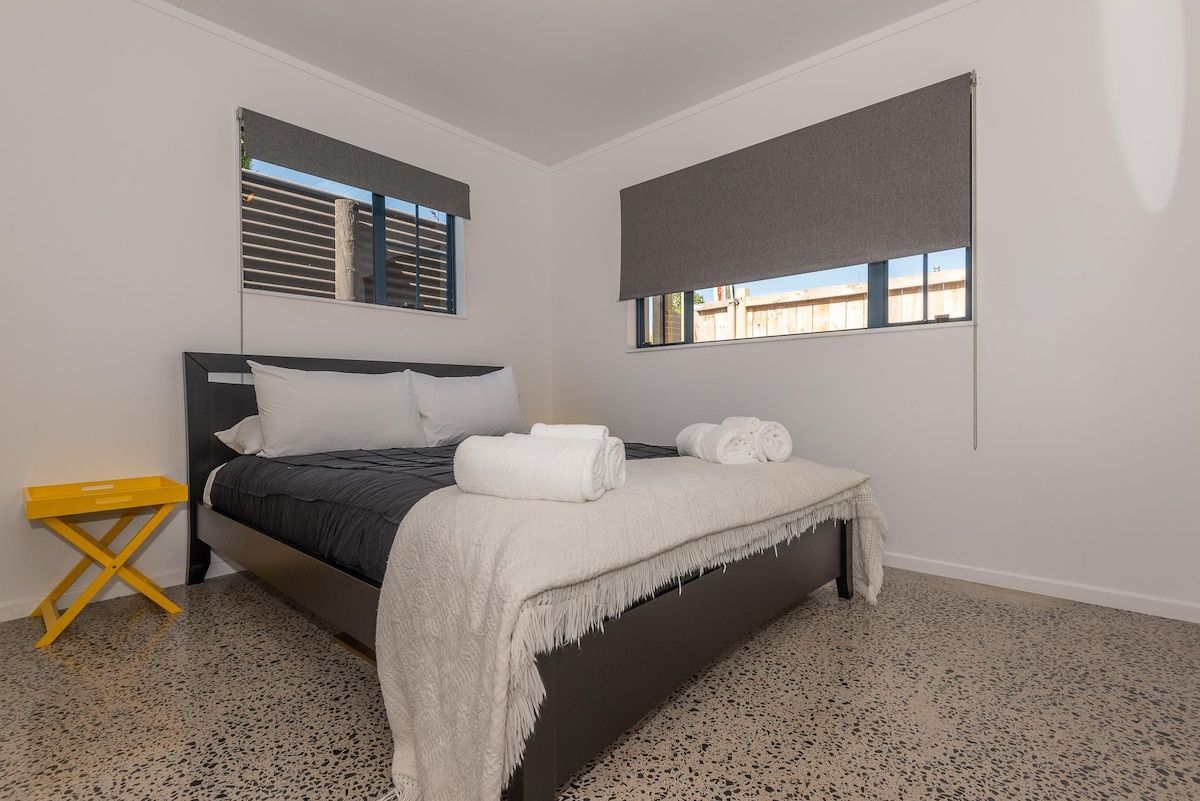 A bedroom with a bed, a small yellow side table, and a window with blinds.