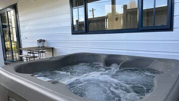 A hot tub outdoors with a table and chairs nearby, a window, and a view of the neighborhood.