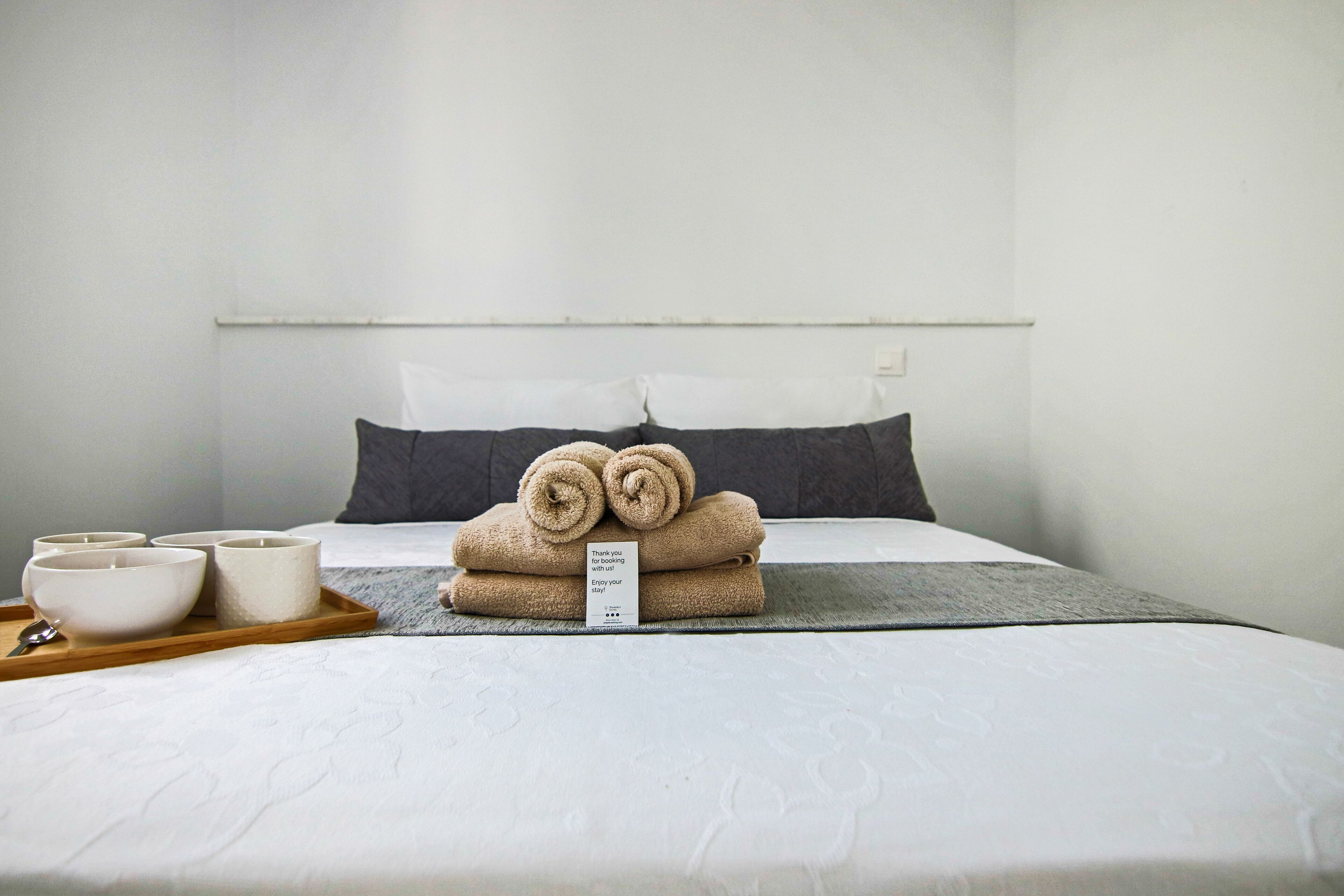 Una cama bien hecha con una bandeja de cuencos, un cartel, un almohadón y una toalla enrollada sobre la cama.