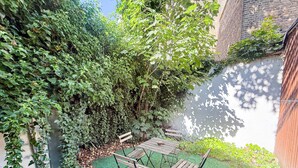 En hyggelig udendørs terrasse med et træbord og to stole, omgivet af frodigt grønt og en murstensvæg.