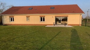 Un bâtiment d’un seul étage, avec un toit recouvert de tuiles rouges, un petit porche doté d’un banc et une pelouse bien entretenue.